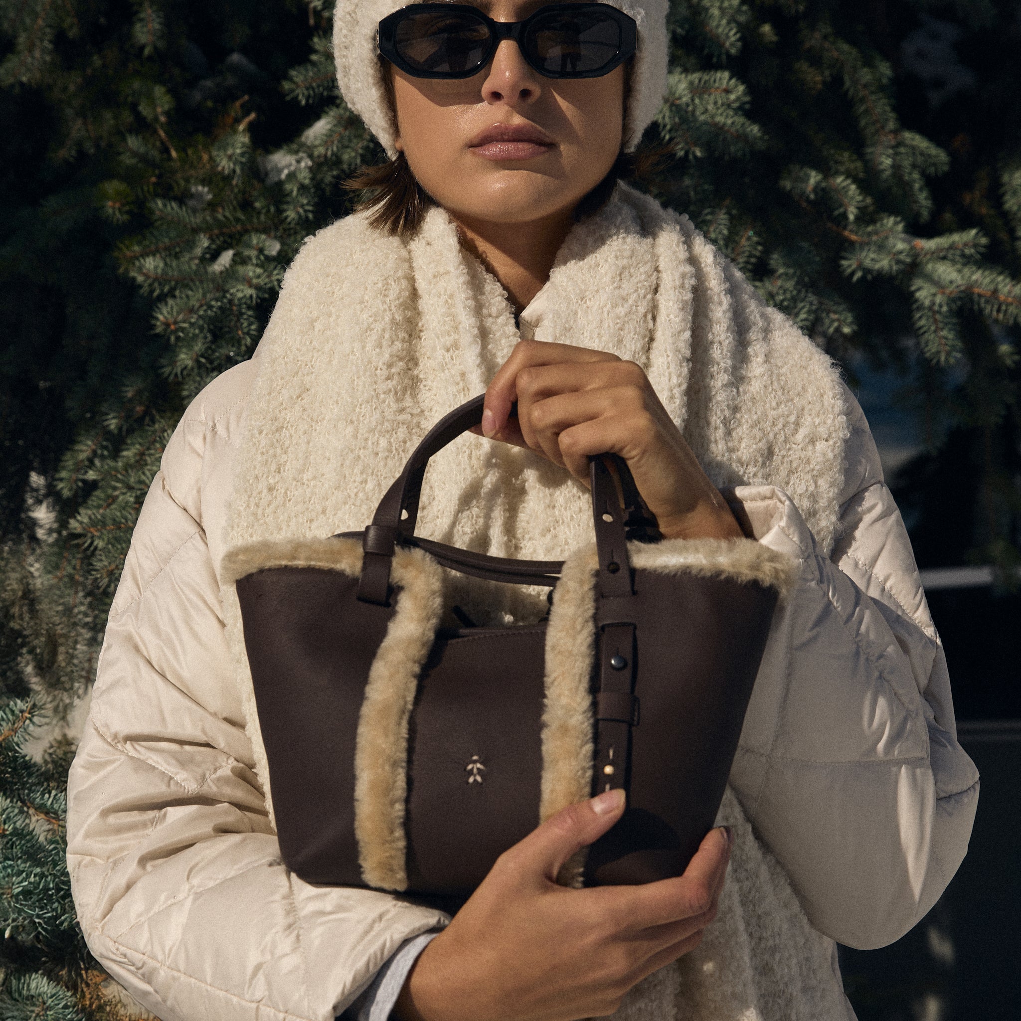 Person wearing a white coat and hat, holding a brown leather bag with fur lining, against a snowy background.