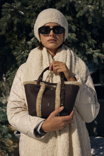 Person wearing a white coat and hat, holding a brown leather bag with fur lining, against a snowy background.