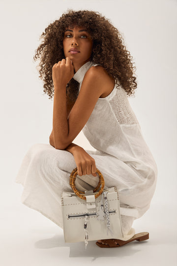 A model with curly brown hair crouches in a white sleeveless dress while holding a structured white handbag with a bamboo ring handle and silver hardware, set against a plain white studio background.