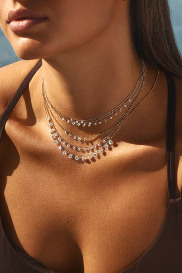 A close-up lifestyle shot of a woman wearing a multi-layered silver necklace with clear crystal-like round beads. The necklace is styled with a dark brown bikini top, set against a warm, sunlit background.