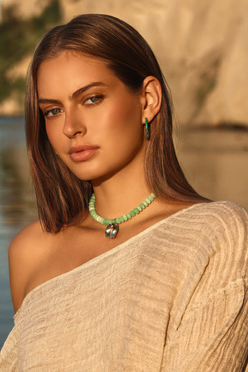A woman wearing a light green beaded necklace with a silver-toned pendant and matching green drop earrings, styled with a beige off-the-shoulder top against a warm, blurred outdoor background.