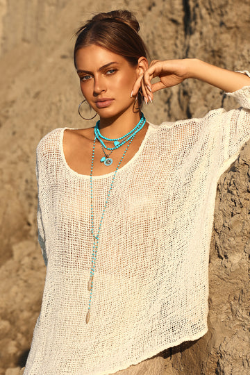 A woman wearing a loose, off-white crochet knit top and layered turquoise necklaces, posing outdoors against a textured stone wall.