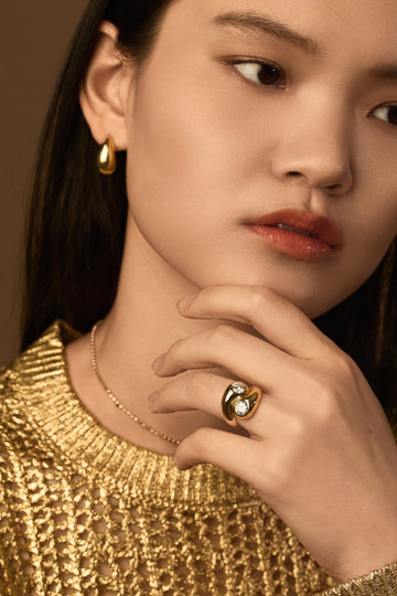 Woman wearing gold jewelry including earrings, necklace, and ring against a brown background