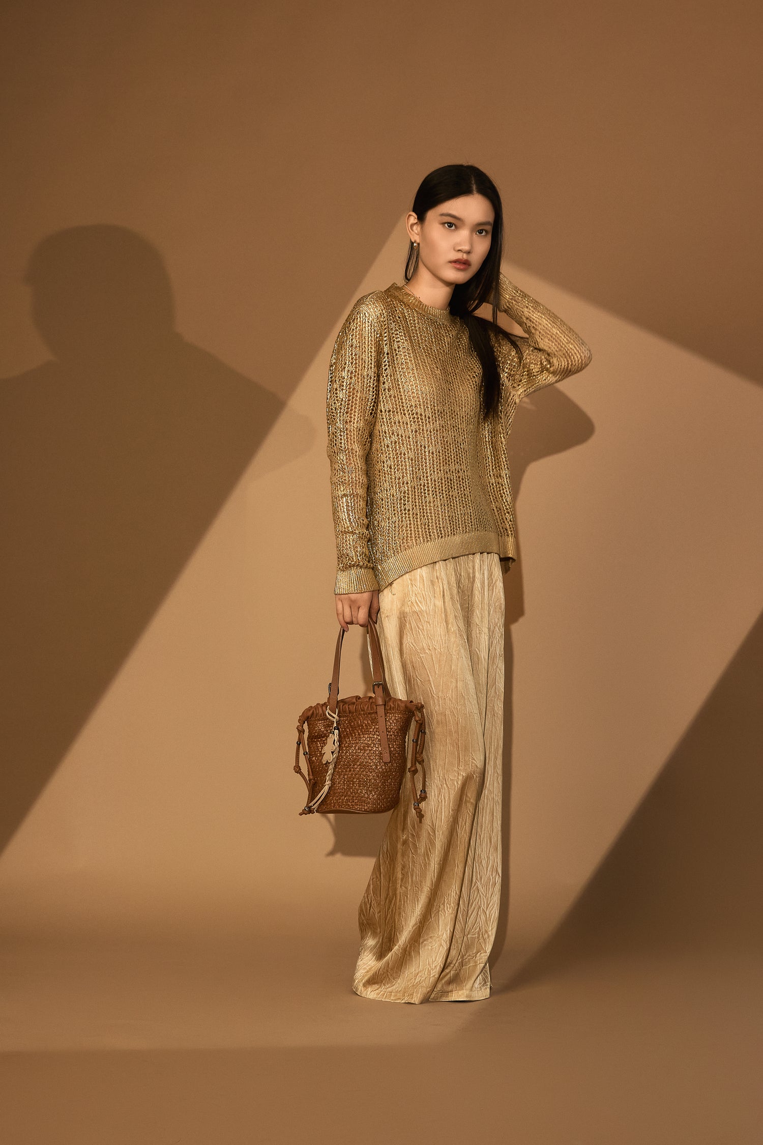 Woman in a hand painted laminated pullover and beige plazzo Touser, holding a brown leather handbag against a brown background