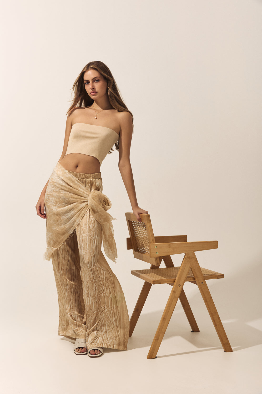 Woman in a beige outfit standing next to a wooden chair on a plain background