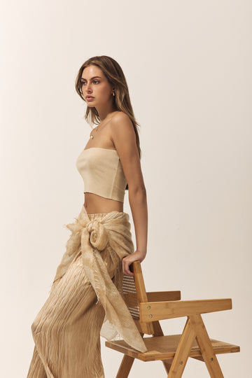 Woman in beige strapless top and long skirt standing next to a wooden chair on a plain background